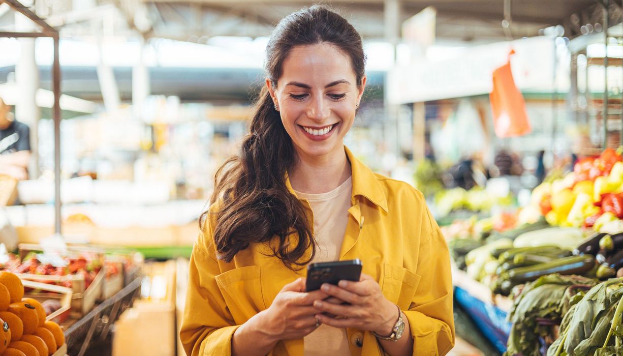 Frau, steht im Supermarkt und schaut auf ihr Smartphone.