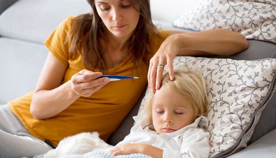 Eine Mutter sitzt auf dem Bett mit ihrem kranken Kind und schaut auf das Fieberthermometer.