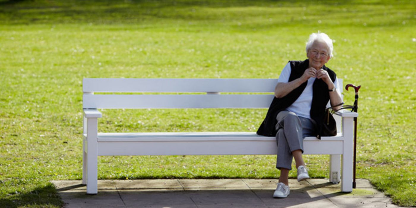 alte Dame sitzt in der Sonne auf einer weißen Parkbank