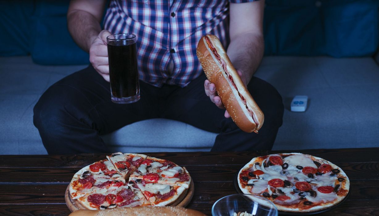 Dicker Mann, sitzt auf einer Couch und isst Pizza, Baguette und trinkt Cola.