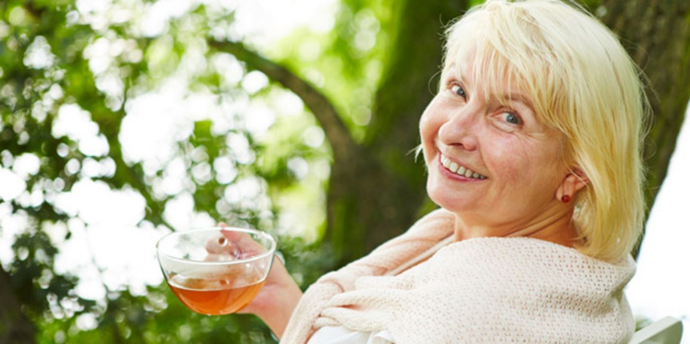 Ältere Frau sitzt in ihrem Garten und hält eine Tasse Tee in der Hand.