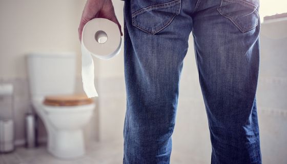 Ein Mann hält eine Rolle Klopapier in der Hand und geht Richtung Toilette (unscharf im Hintergrund erkennbar).