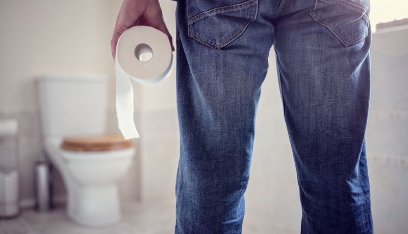 Ein Mann hält eine Rolle Klopapier in der Hand und geht Richtung Toilette (unscharf im Hintergrund erkennbar).