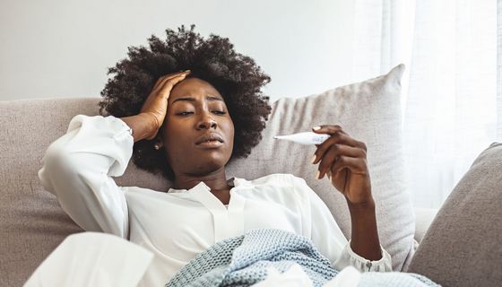 Eine junge Frau liegt in einer Decke auf dem Sofa, greift sich an den Kopf und blickt auf ein Fieberthermometer.