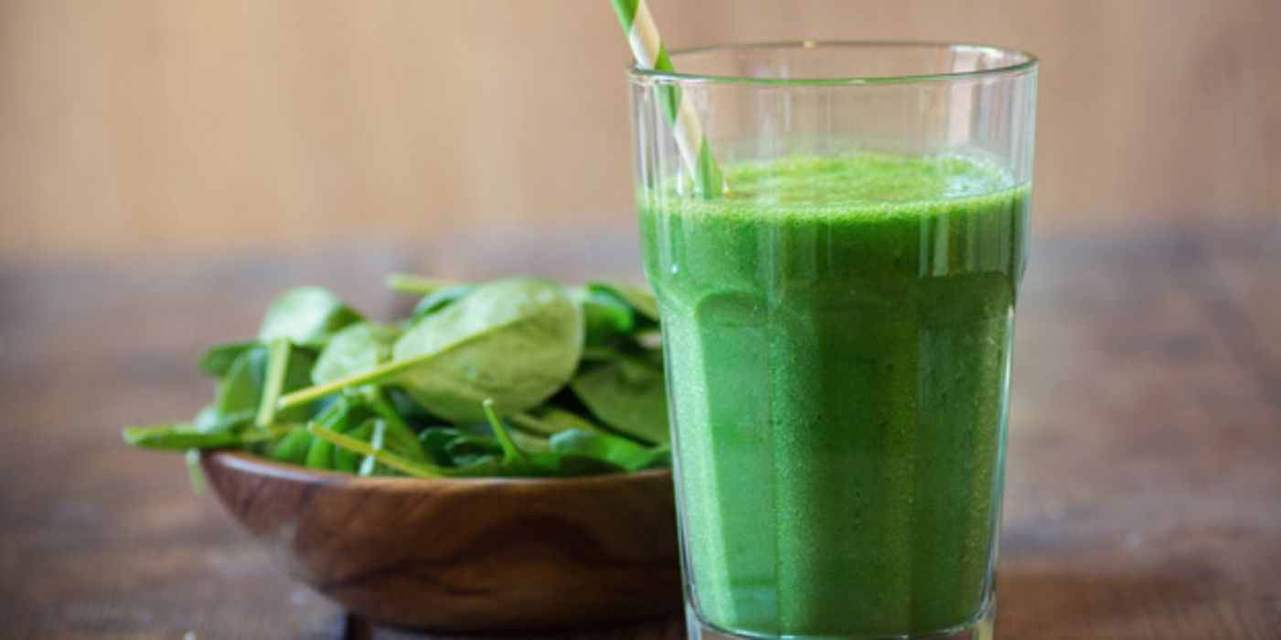 Spinatdrink in hohem Glas mit weiß-grünem Strohhalm auf dunkelm Holztisch, links etwas dahinter dunkles Holzschälchen mit jungen Spinatblättern