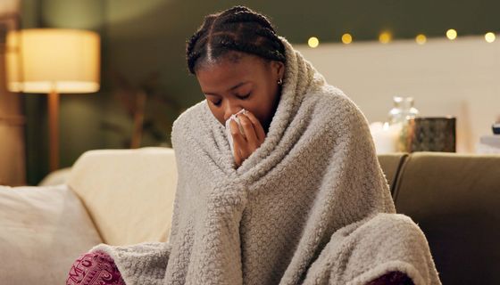 Eine junge Frau sitzt auf einem Sofa in eine Decke gewickelt. Sie schnäuzt sich die Nase.