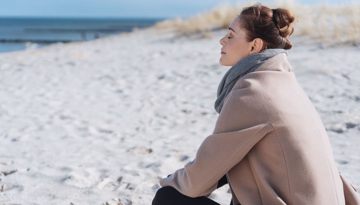 Junge Frau, sitzt an einem kühleren Tag warm angezogen am Strand und sonnt sich.