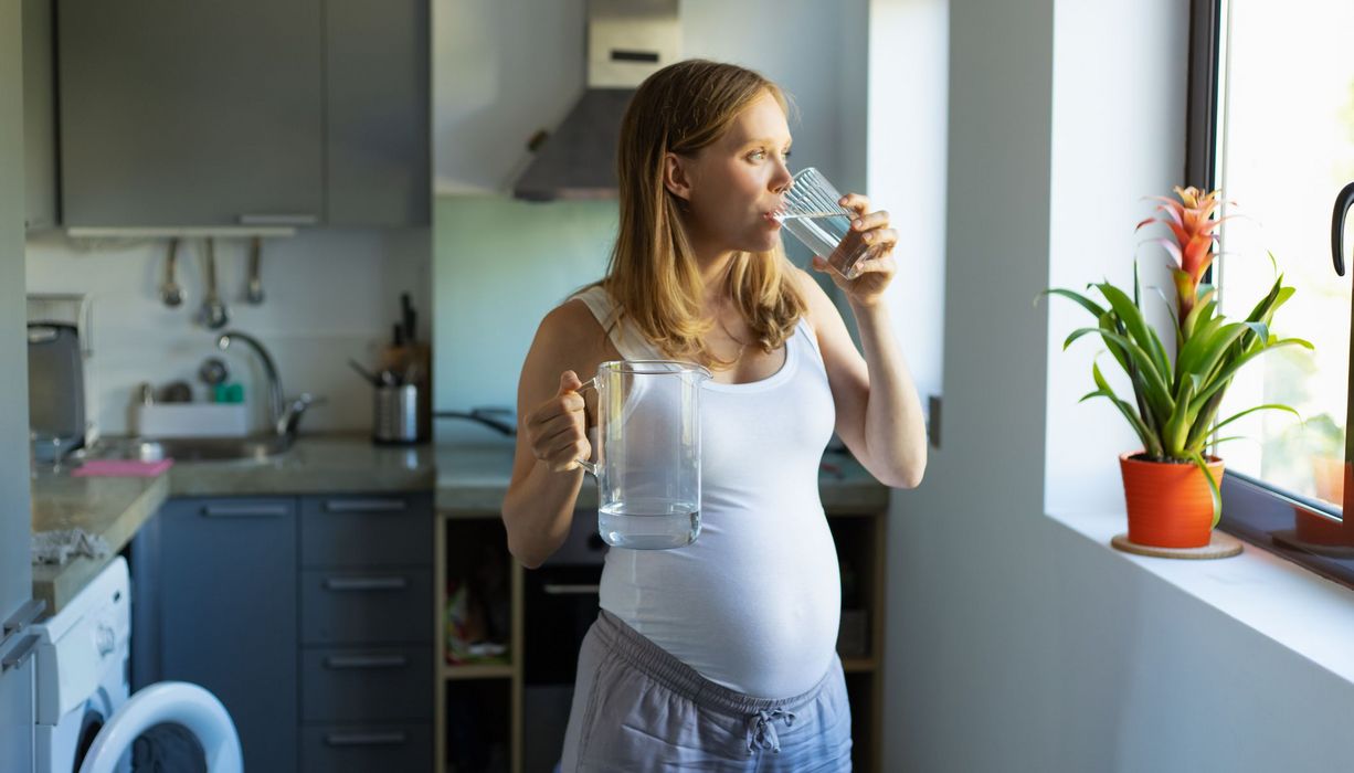 Junge schwangere Frau, trinkt Wasser in der Küche und schaut aus dem Fenster.