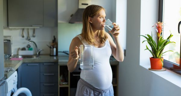 Junge schwangere Frau, trinkt Wasser in der Küche und schaut aus dem Fenster.