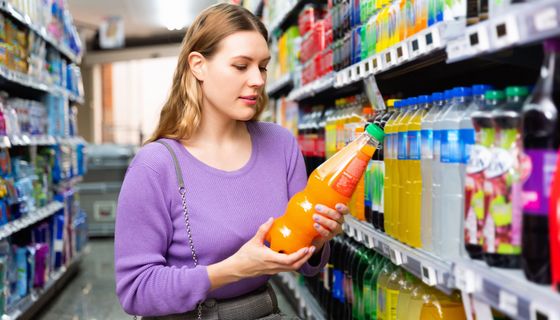 Eine Frau steht vor dem Getränkeregal und schaut auf die Zutatenliste eines Softdrinks.