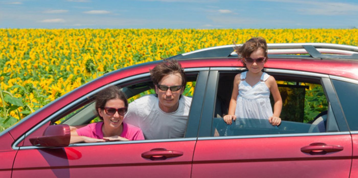 Mutter, Vater, Tochter schauen lachend aus Fenstern eines roten Autos, im Hintergrund Sonnenblumen und blauer Himmel