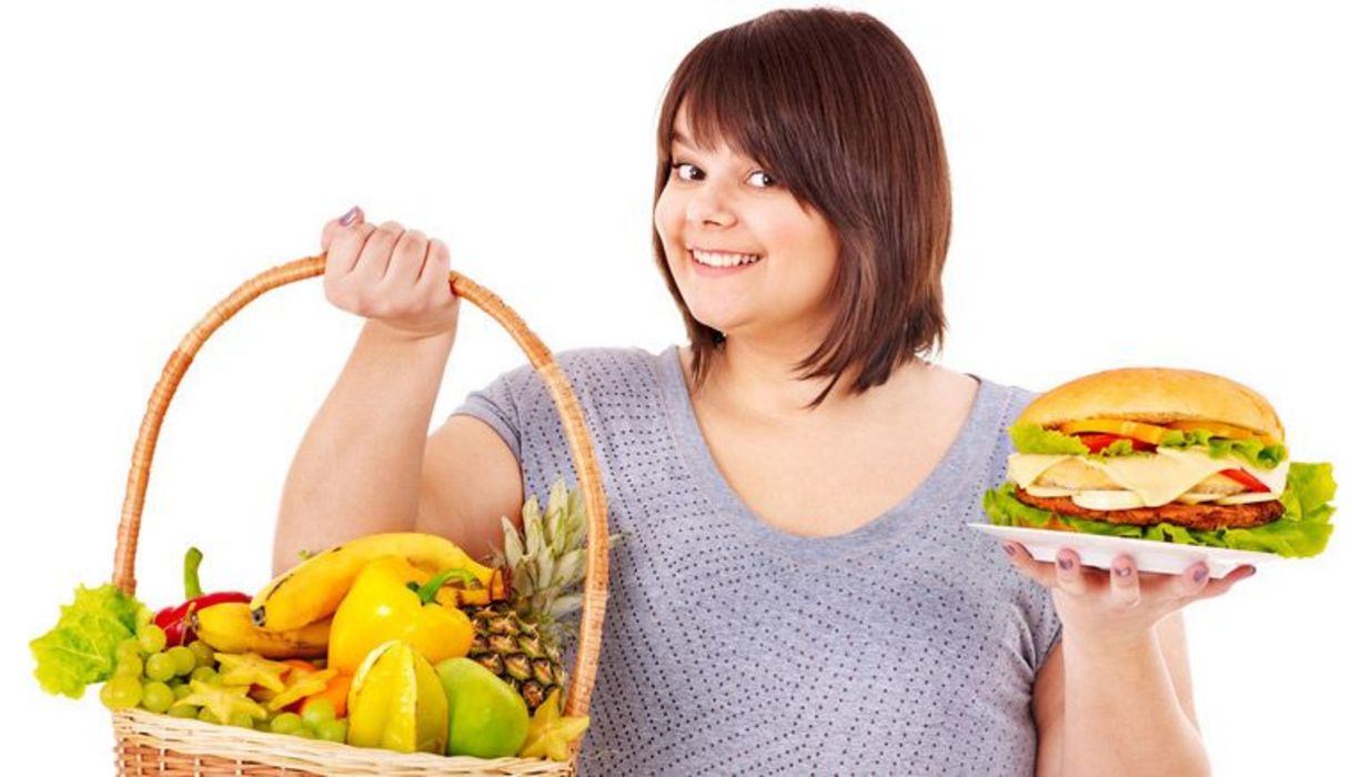 Mollige junge Frau mit einem Obstkorb in der einen und einem Sandwich in der anderen Hand