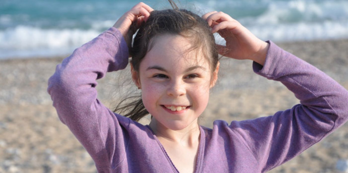 Mädchen, ca. 9 Jahre, Meer im Hintergrund, lila Feinstrickjacke, fasst sich ins Haar und blinzelt in die Kamera