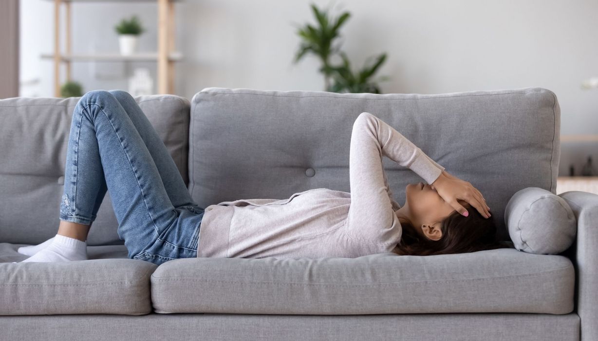 Junge Frau, liegt auf der Couch, hält ihre Hände an den Kopf.