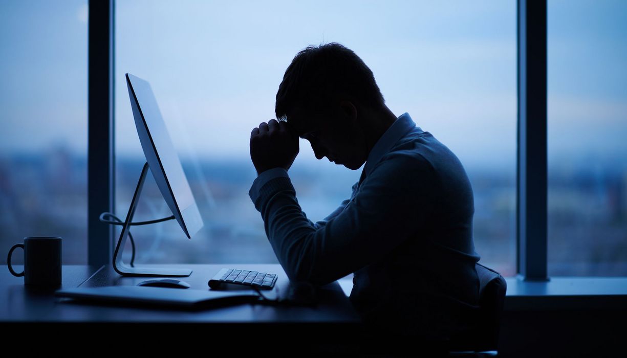 Mann, sitzt vor dem Computer im Büro und stützt den Kopf in die Hände.