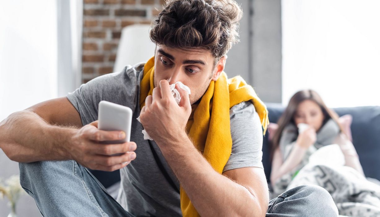 Junger Mann, putzt sich die Nase und schaut auf sein Handy.