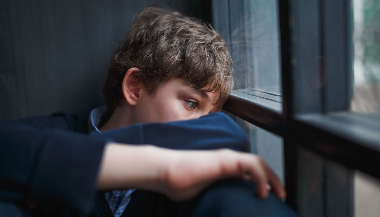 Trauriger Junge mit leerem Blick sitzt am Fenster.