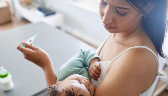 Junge Mutter, hält ihr Baby im Arm und ein Fieberthermometer in der Hand.