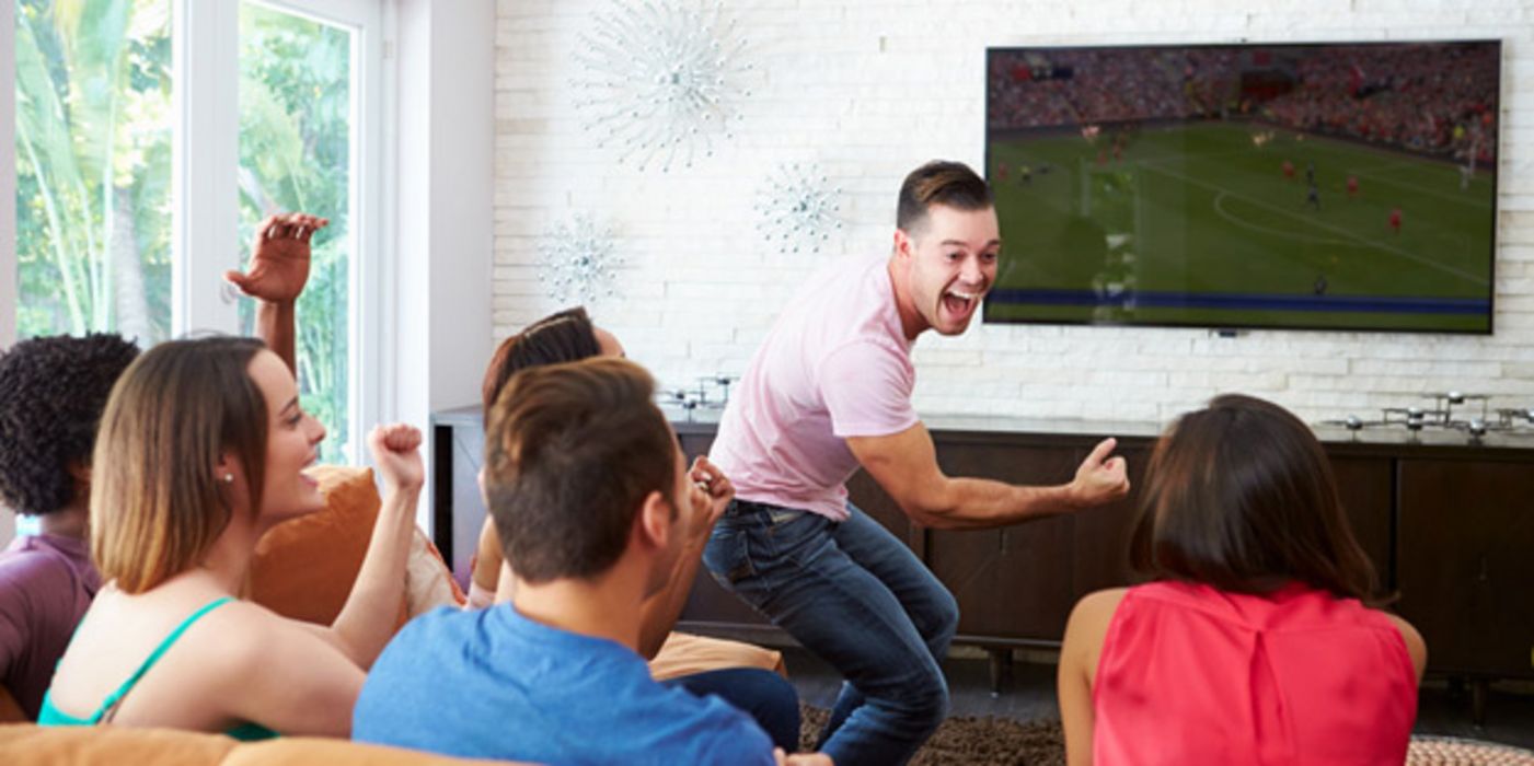 Gruppe junger Menschen schaut im Wohnzimmer ein Fußballspiel im Fernsehen, ein junger Mann steht, ballt die Faust und feuert seine Mannschaft an