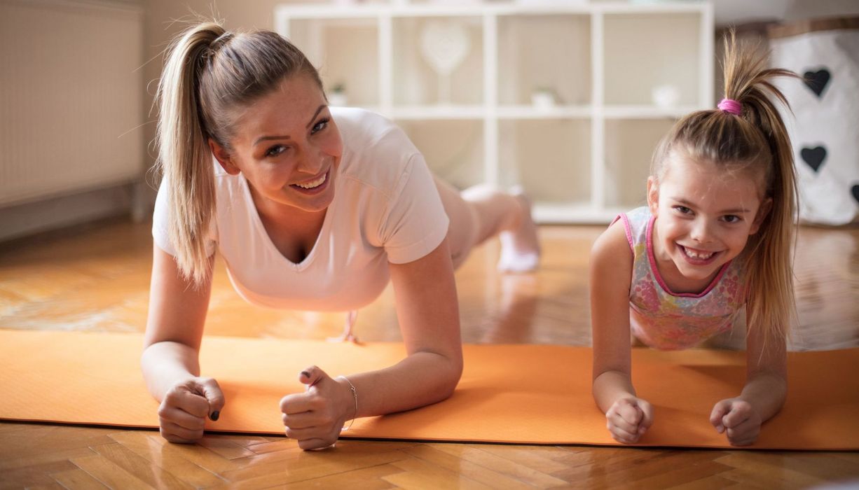 Junge Frau und Tochter machen zusammen im Wohnzimmer Sport.