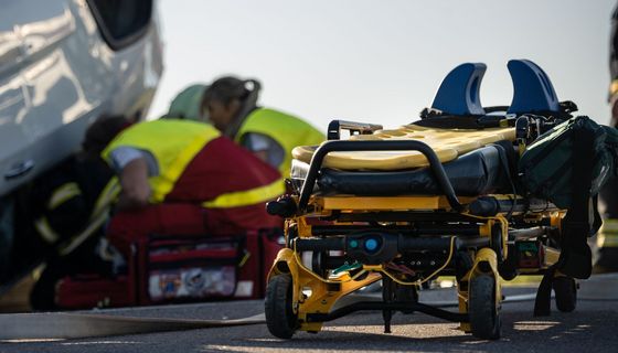 Im Vordergrund: Eine spezielle Liege mit Stütze für den Nacken, im Hintergrund: Ein Unfallwagen auf dem Dach, Sanitäter und Feuerwehr in der Hocke vor dem Fenster.