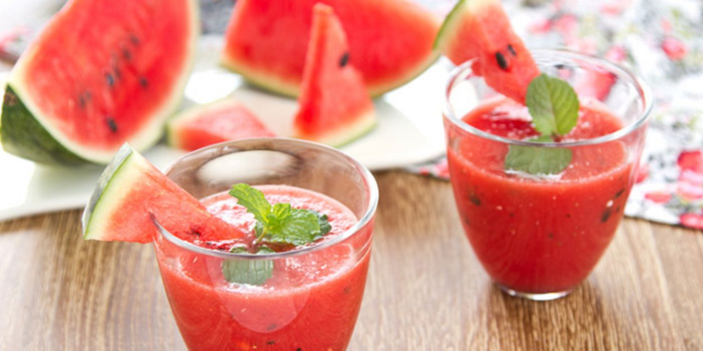 Wassermelonensaft in 2 Gläsern, dahinter aufgeschnittene Wassermelonen