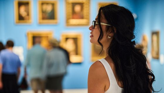 Im Vordergrund: Eine junge Frau, die nach links aus dem Bild heraus blickt, im Hintergrund (unscharf): Blaue Wände mit historischen Portraits und eine kleine Gruppe Museumsbesucher.