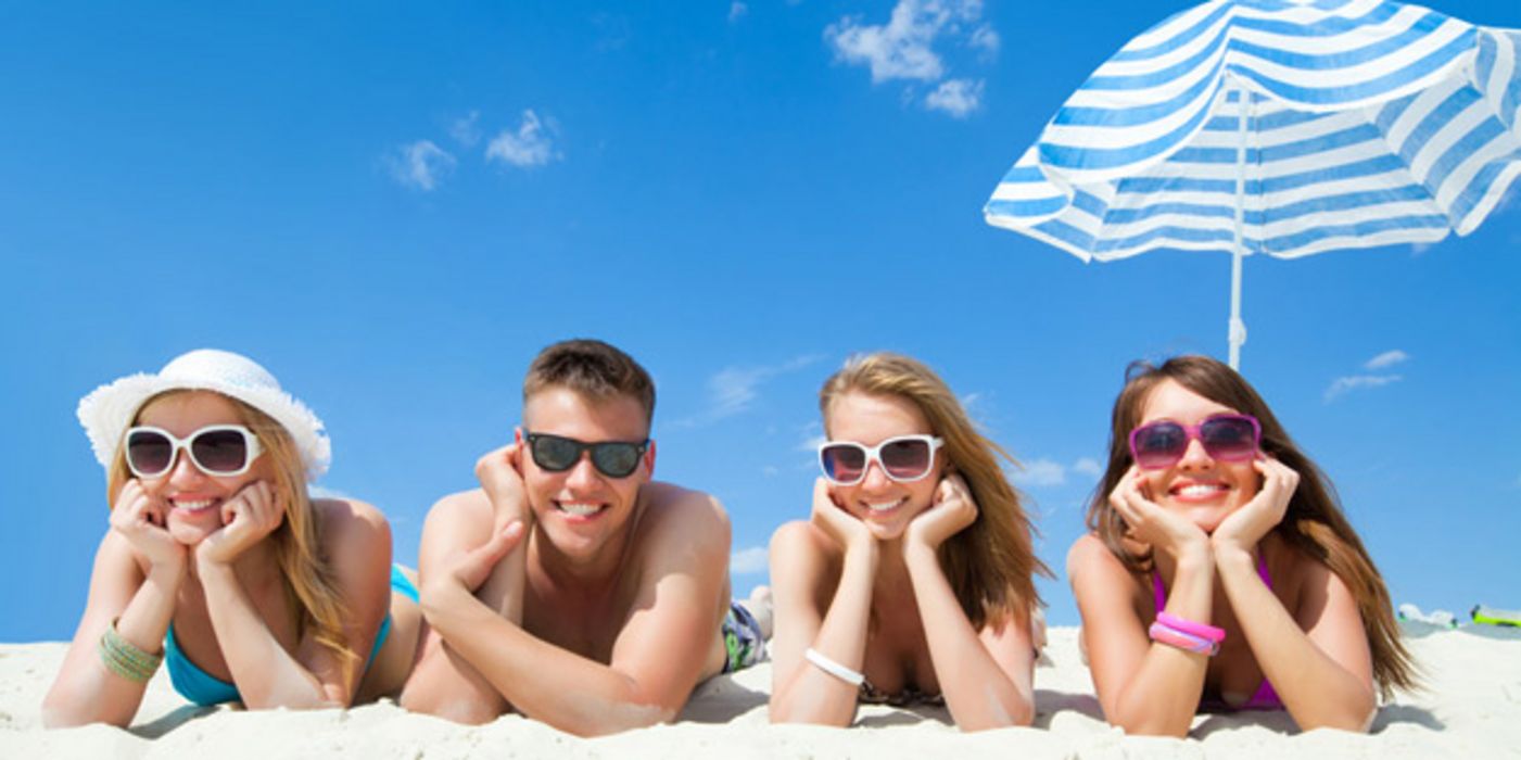 Vier junge Erwachsene liegen am Strand auf dem Bauch und tragen Sonnenbrillen, im Hintergrund ein Sonnenschirm