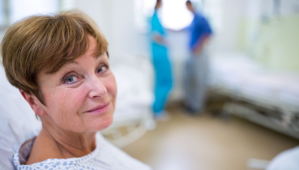 Frau, etwa 50 Jahre alt, sitzt im Krankenhausbett und lächelt.