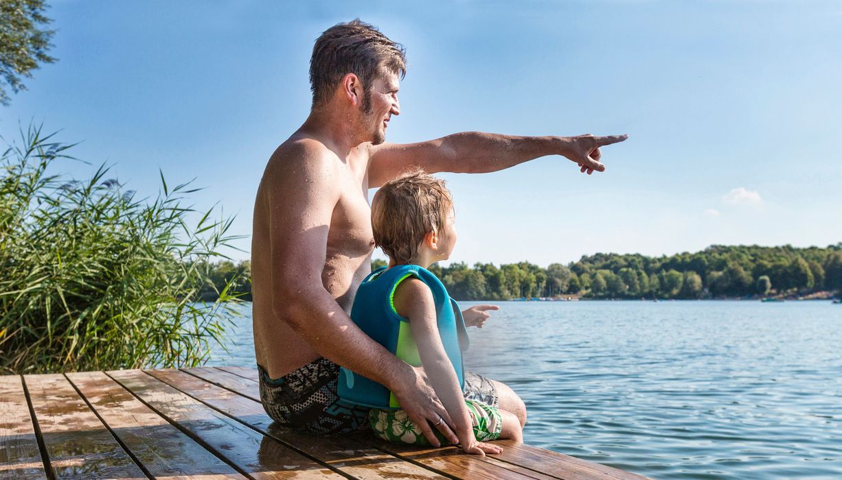 Vater, sitzt mit seinem Kind an einem Badesee.