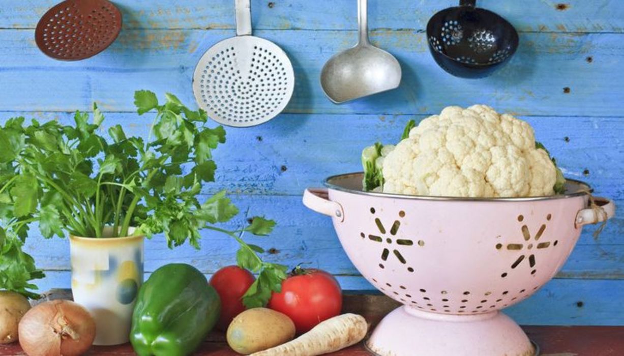 Blumenkohl in weißem, altmodischem Metallsieb, daneben Tomaten, grüne Paprika, Petersilie in irdenem Gefäß: Stillleben vor hellblau gestrichener Holzwand auf rotbraun abgeblättertem Holz