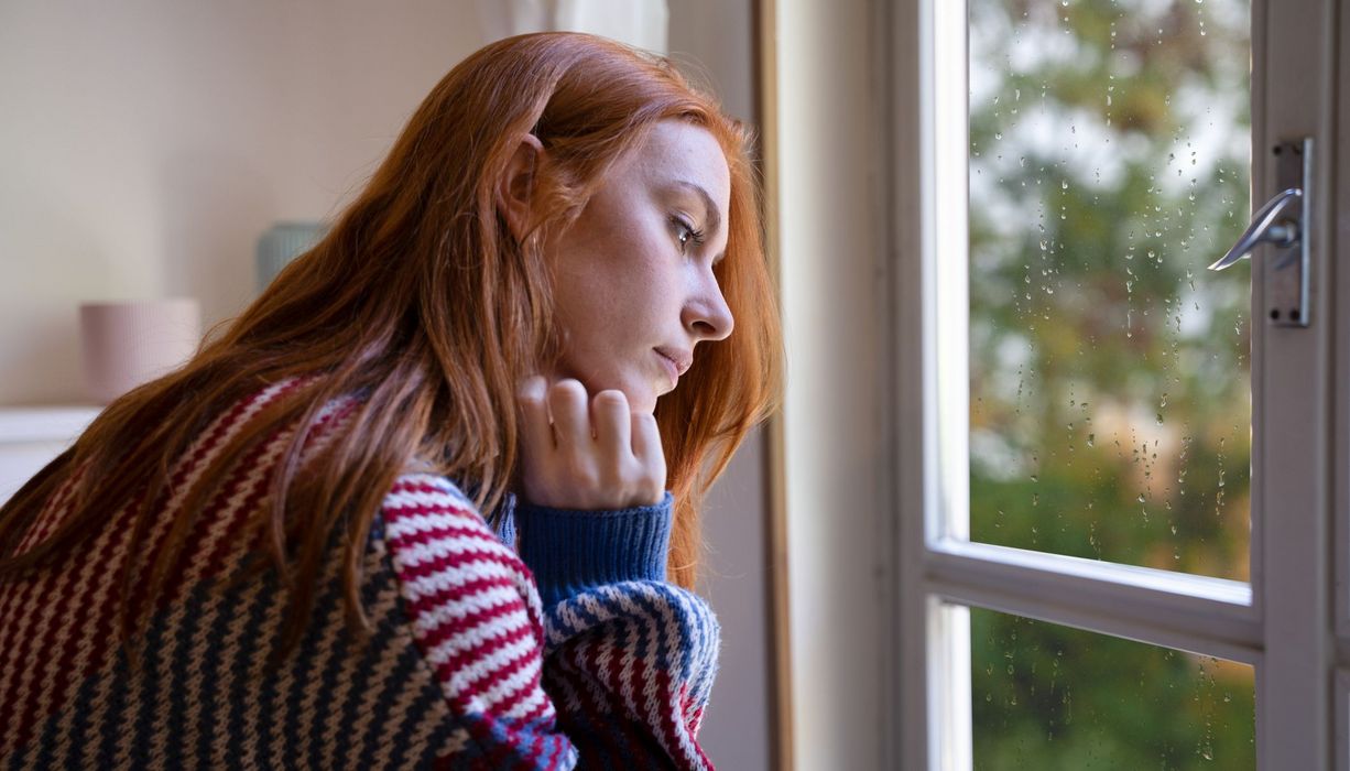 Junge Frau, sitzt vor dem Fenster und schaut traurig nach unten.