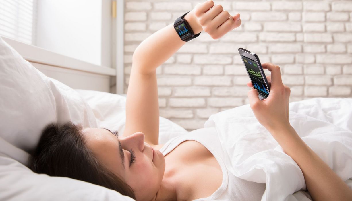 Junge Frau, liegt im Bett und überprüft mit einer Smartwatch ihren Puls.