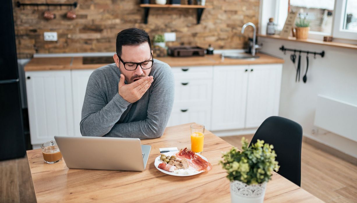 Junger Mann, sitzt mit seinem Laptop und einem Teller mit Essen in der Küche und gähnt.