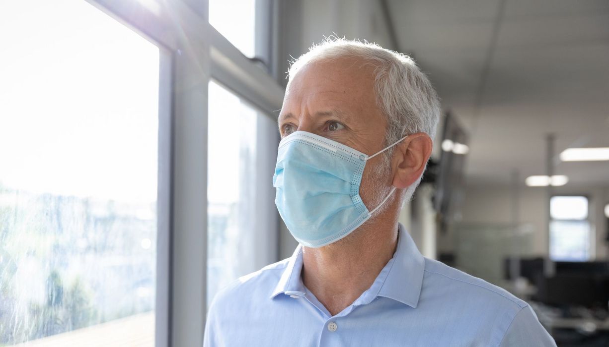 Älterer Mann mit Maske blickt aus dem Fenster.