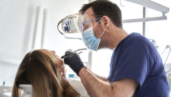 Zahnarzt behandelt eine Patientin, in beiden Händen hält er Werkzeuge. Ihr Gesicht ist nicht zu sehen.