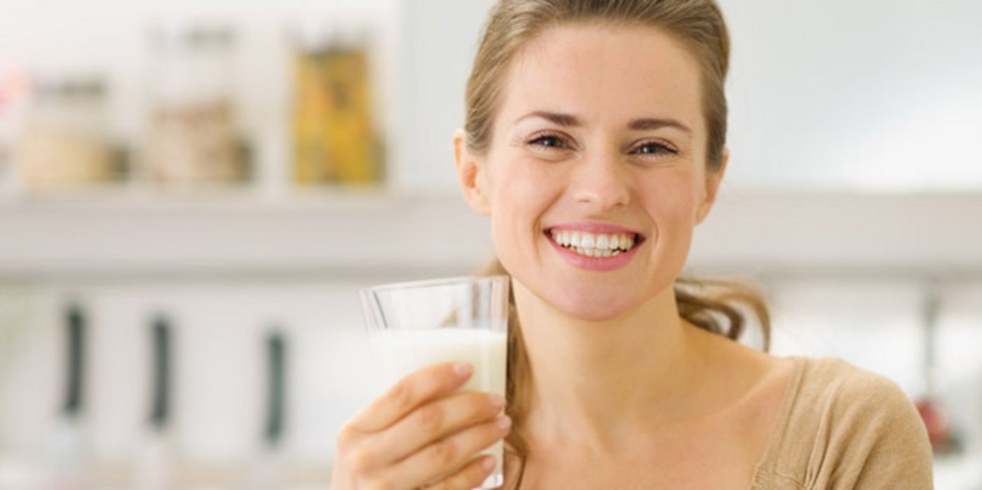 Junge, in die Kamera lachende Frau, die langen Haare zurückgebunden, mit einem Glas Milch in der linken Hand