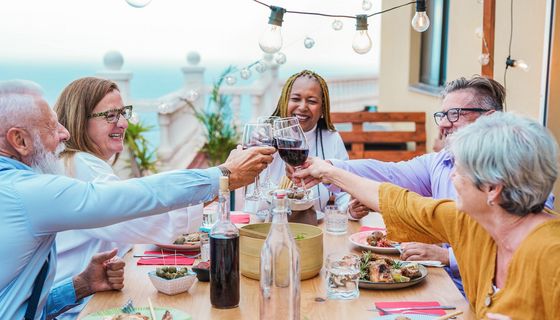 Freunde im mittleren Alter sitzen an einem gedeckten Tisch und prosten sich mit Rotwein zu. zu.