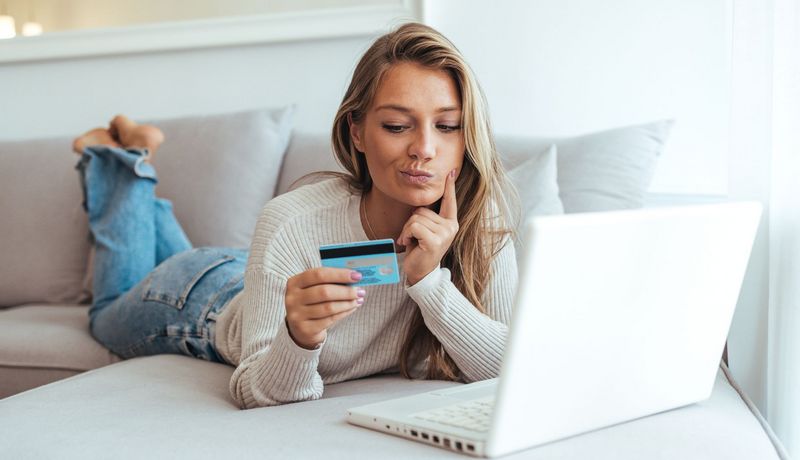 Junge Frau schaut zweifelnd auf ihre Kreditkarte, vor ihr steht ihr aufgeklappter Laptop.