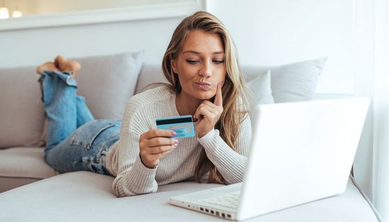 Junge Frau schaut zweifelnd auf ihre Kreditkarte, vor ihr steht ihr aufgeklappter Laptop.