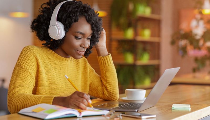 Junge Frau mit Kopfhörern sitzt lächelnd vor ihrem Laptop und schreibt etwas in ein Notizbuch.