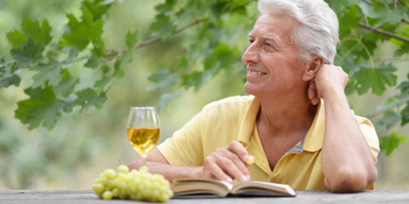 Porträtfoto weißhaariger Mann, gelbes Poloshirt, linker Elenbogen auf Tisch gestützt, Hand am Gesicht, Weißwein und Buch vor sich, Laubbaum hinter sich, blickt leicht nach oben ins Off