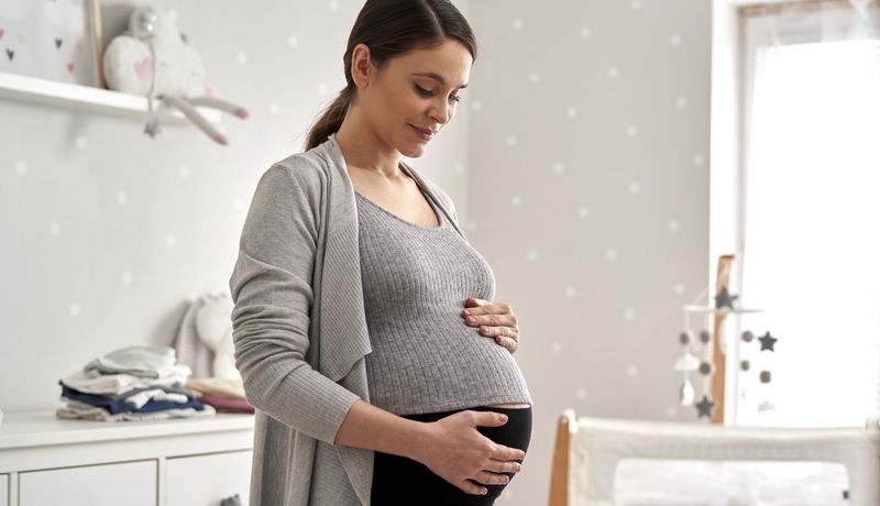 Eine hochschwangere Frau streichelt sich im Babyszimmer den Bauch und lächelt.
