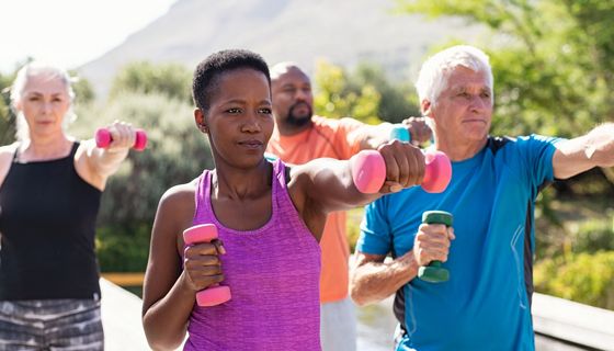 Vier Erwachsene unterschiedlichen Alters, zwei Frauen, zwei Männer, trainieren unter freiem Himmel mit Hanteln.