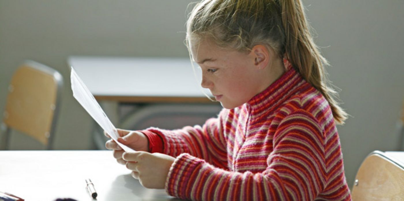 Schülerin, ca. 8 Jahre, Profilbild, Pferdeschwanz und rot geringelter Pulli, liest konzentrirt im Klassenraum von einem Blatt