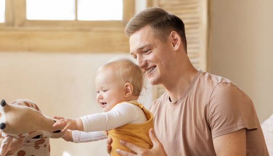 Junge Familie: Der Vater hält das etwa 10 Monate alte Kind, das versucht auf seinen Füßen zu stehen und nach einem Kuscheltier greift.