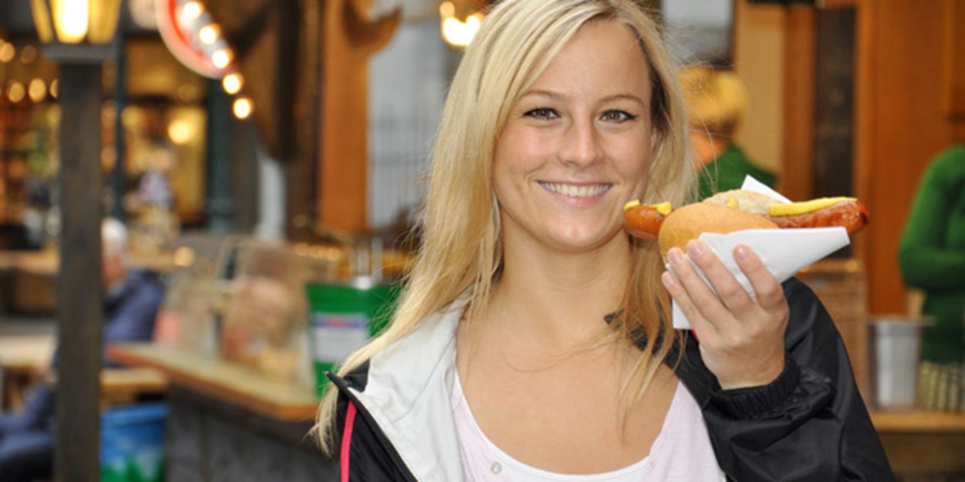 Junge blonde Frau abends vor einer Imbissbude, in der Hand eine Rostbratwurst im Brötchen mit Senf