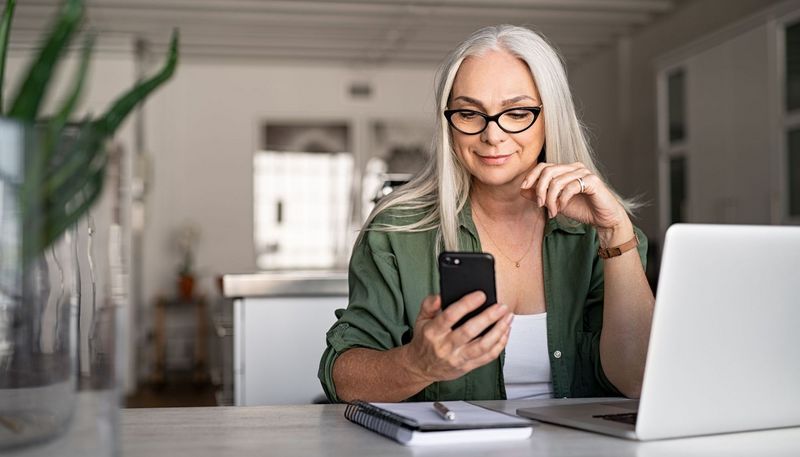 Ältere Frau, ca. 60 Jahre alt, sitzt am Schreibtisch vor einem Laptop mit einem Telefon in der Hand.