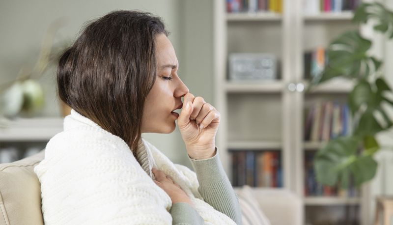 Junge Frau, sitzt auf einer Couch und hustet.