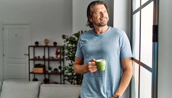 Ein Mann schaut aus dem Wohnzimmerfenster und trinkt einen Kaffee.